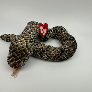 May include: A brown and black snake plush toy with a gold tongue. The toy has a Ty Beanie Babies tag attached to it.
