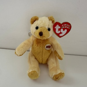 Peut inclure: Un petit ours en peluche beige avec un nez brun et des yeux noirs. L'ours est assis les jambes croisées et a une étiquette Ty attachée à son oreille droite. L'étiquette est rouge avec du texte blanc qui dit "Ty" et "Jiggle Beanies".
