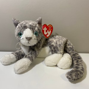 May include: A gray and white plush cat with blue eyes and a red Ty Beanie Baby tag. The cat is lying on its side with its head turned to the side.