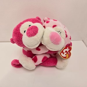 Peut inclure: Deux peluches Beanie Babies roses se font un câlin. Les Beanie Babies portent des motifs de cœurs roses et blancs. L'un des Beanie Babies a un nez rose et l'autre un nez blanc. Les Beanie Babies ont des yeux noirs. Le Beanie Baby de droite a une étiquette Ty attachée à son bras.