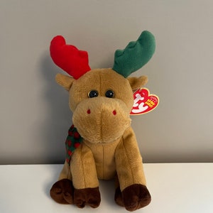 May include: A brown plush moose with red and green antlers. The moose is wearing a green and red scarf with white polka dots. The moose is sitting on a white surface. The moose has a red and white "Ty" tag attached to its ear. The tag says "Original Beanie Babies".