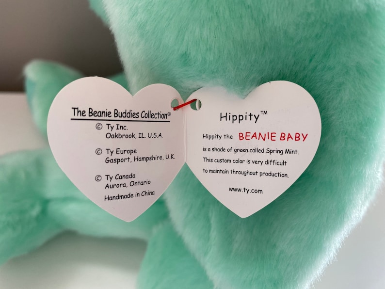 Puede incluir: Un peluche verde menta con una etiqueta en forma de coraz&oacute;n. La etiqueta dice "Hippity the BEANIE BABY" y menciona el color "Spring Mint". La etiqueta tambi&eacute;n incluye el texto "The Beanie Buddies Collection" y la ubicaci&oacute;n del fabricante.