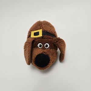 May include: Brown plush toy dog with a black nose, white eyes, and a yellow and black belt around its head.