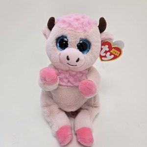 May include: Pink plush cow with large blue eyes and a brown nose. The cow is sitting with its legs crossed and has a pink and white fur pattern. The cow has a Ty Beanie Babies tag attached to its ear.