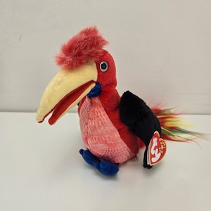 May include: A plush bird toy with a red body, black wings, and a yellow beak. The bird has a red crest and blue feet. A tag is attached to the bird's wing.