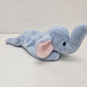May include: A light blue plush elephant toy with pink ears. The elephant is lying on its side with its trunk raised.