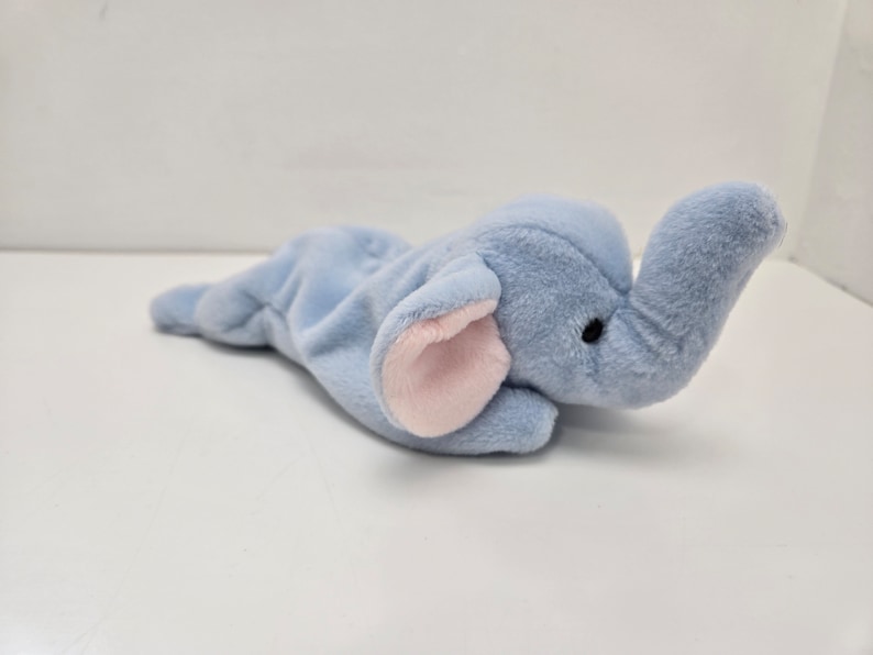 May include: A light blue plush elephant toy with pink ears. The elephant is lying on its side with its trunk raised.