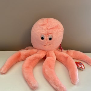 May include: A plush pink octopus toy with eight long tentacles and a smiling face. The toy has large, round eyes and a tag attached to one of its tentacles. The octopus is made of soft, fuzzy material.