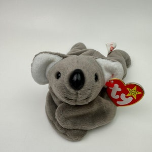 May include: A gray and white plush koala bear with black eyes and a black nose. The bear is lying on its stomach with its arms outstretched. The bear has a red and white Ty Beanie Baby tag attached to its right arm.