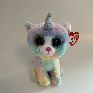 Puede incluir: Un gato de peluche de color arcoíris pastel con un cuerno de unicornio de purpurina plateada, ojos grandes azules y dorados y bigotes rosas. El gato está sentado sobre un fondo blanco con una etiqueta roja de Ty Beanie Boo's.