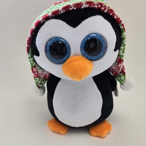 May include: A plush penguin toy wearing a red and green knit hat with a white pom-pom. The penguin has large blue eyes and a white belly.