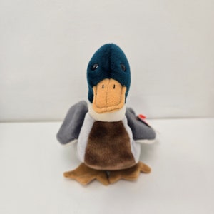 Pu&ograve; includere: Un'anatra di peluche blu e marrone con un becco bianco e occhi neri. L'anatra &egrave; seduta su una superficie bianca.