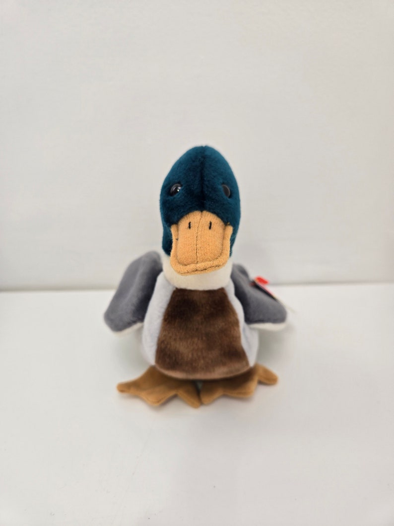 Pu&ograve; includere: Un'anatra di peluche blu e marrone con un becco bianco e occhi neri. L'anatra &egrave; seduta su una superficie bianca.