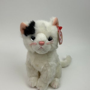 Pode incluir: Um gato de pelúcia branco com uma mancha preta sobre um olho e detalhes rosa no nariz, orelhas e patas. Possui uma etiqueta vermelha anexada. O gato está sentado, voltado para a frente.