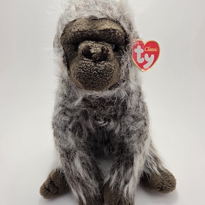 May include: A plush gorilla toy with dark brown textured fabric on the face, hands, and feet, covered in long, shaggy grey and white fur. A heart-shaped red tag with the white text 'ty' and 'Classic' is attached to the left side of the head.