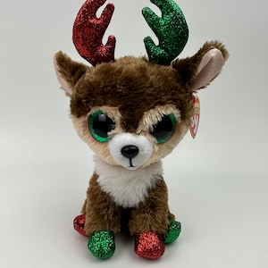 May include: A plush reindeer toy with brown fur, large green eyes, and a white chest. The antlers are red and green glitter. The reindeer is sitting with its front legs forward and its back legs tucked under.