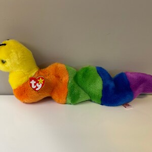 Op de afbeelding: Een gele en regenboog gestreepte Ty Beanie Baby worm knuffel met een rood Ty-label waarop "Ty" staat en een ster.