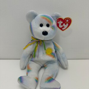 Könnte beinhalten: Ein weißer Plüsch-Regenbogenbär mit einer gelben Sonne auf der Brust. Der Bär hat ein rotes Beanie Baby-Etikett am Ohr, auf dem "Beanie Baby" steht und ein rotes "ty"-Logo.