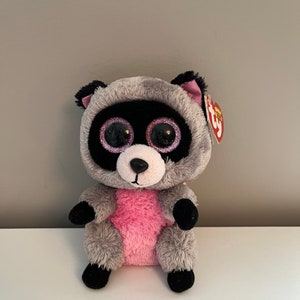May include: A gray and pink raccoon plush toy with large glittery eyes. The toy has a pink belly and black feet. The toy has a Ty tag attached to its ear.