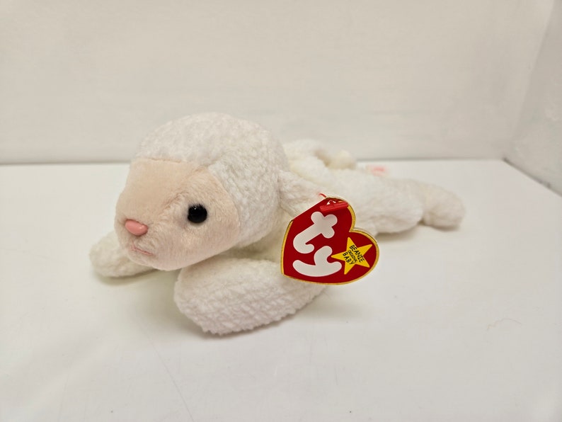 May include: A white plush lamb toy with a pink nose and ears, and black eyes. The lamb is lying down with its legs extended. A red heart-shaped tag is attached to the lamb.