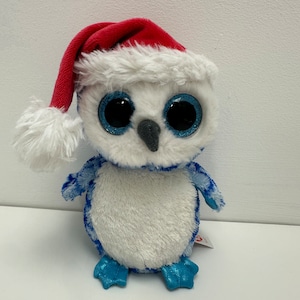 May include: White and blue plush owl wearing a red Santa hat with white fur trim. The owl has large blue eyes with glitter.