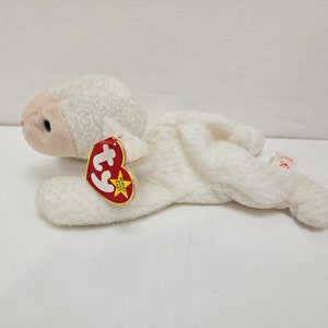 May include: A white plush lamb toy with a red and yellow tag. The lamb is lying down with its head and front legs raised. The tag has the word "BEANIE BABY" on it.