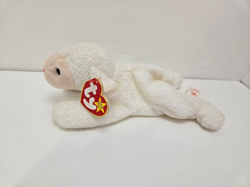 May include: A white plush lamb toy with a red and yellow tag. The lamb is lying down with its head and front legs raised. The tag has the word "BEANIE BABY" on it.