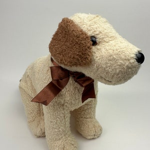 Op de afbeelding: Een pluche wit honden speelgoed met bruine oren en een bruine strik. De hond zit met zijn hoofd schuin.