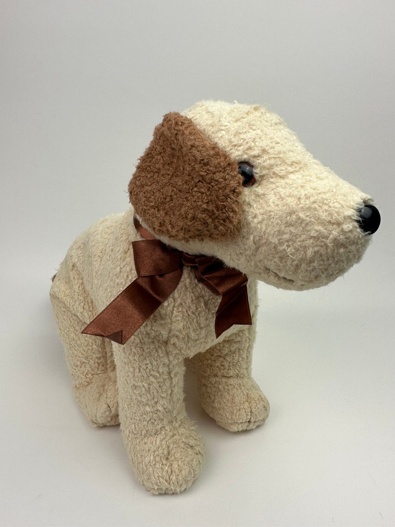 Op de afbeelding: Een pluche wit honden speelgoed met bruine oren en een bruine strik. De hond zit met zijn hoofd schuin.