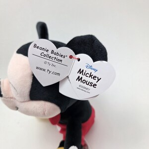 May include: A Mickey Mouse Beanie Baby plush toy. The plush has a black head and ears, a white face, and a red bow tie. The toy has a white heart-shaped tag that reads "Beanie Babies Collection" and another tag that reads "Disney Mickey Mouse".
