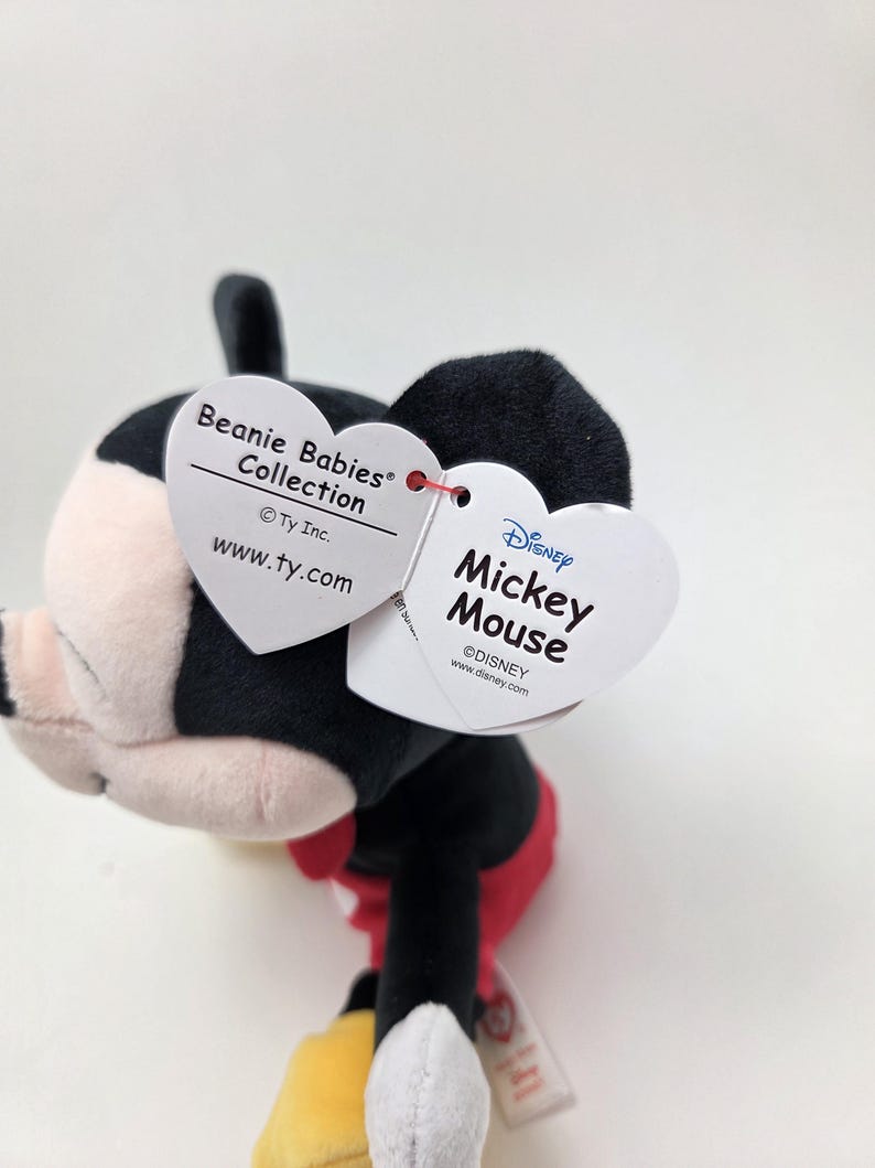 May include: A Mickey Mouse Beanie Baby plush toy. The plush has a black head and ears, a white face, and a red bow tie. The toy has a white heart-shaped tag that reads "Beanie Babies Collection" and another tag that reads "Disney Mickey Mouse".