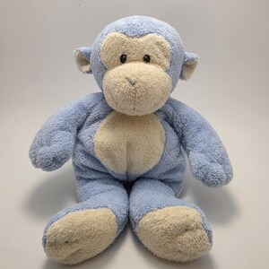 May include: A plush monkey toy in light blue and cream colors. The stuffed animal has a light blue body, head, arms, and legs, with cream-colored accents on the face, hands, and feet. The monkey is sitting upright.