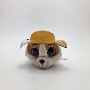 May include: A soft toy dog with big, round eyes, brown and white fur, and a yellow hat. The dog has brown ears and a small pink tail. The toy is designed to look like a puppy.