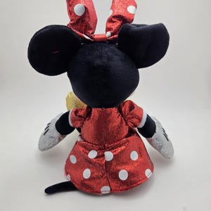 May include: A plush Minnie Mouse doll, seen from the rear, with a red dress and white polka dots, and a matching bow. The doll has black ears, a black tail, and silver gloves. The design is a classic representation of the character.