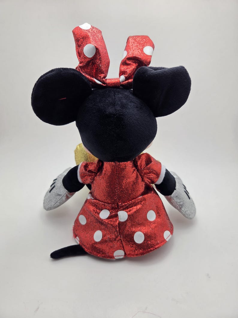May include: A plush Minnie Mouse doll, seen from the rear, with a red dress and white polka dots, and a matching bow. The doll has black ears, a black tail, and silver gloves. The design is a classic representation of the character.