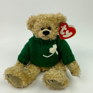 May include: A plush brown teddy bear wearing a green knitted sweater with a white shamrock design. The bear has a red Ty tag attached to its ear.