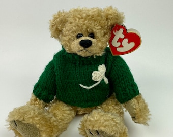 Peut inclure: Un ours en peluche brun portant un pull en tricot vert avec un trèfle blanc. L'ours a une étiquette Ty rouge attachée à son oreille.