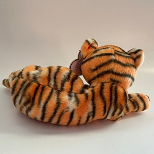 Op de afbeelding: Een pluche tijger knuffeldier met oranje en zwarte strepen. De tijger ligt op zijn zij met zijn kop naar de zijkant gedraaid. De tijger heeft een rood hartvormig label aan zijn oor.