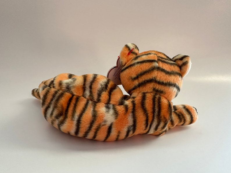 Op de afbeelding: Een pluche tijger knuffeldier met oranje en zwarte strepen. De tijger ligt op zijn zij met zijn kop naar de zijkant gedraaid. De tijger heeft een rood hartvormig label aan zijn oor.