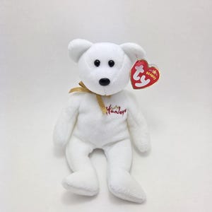 May include: White plush beanie baby bear with a gold ribbon around its neck. The bear has a red embroidered heart on its chest with the word "Hamleys" in white lettering. The bear has a red Ty tag attached to its right ear.