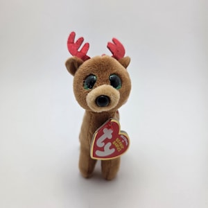 May include: A brown plush reindeer toy with large, glittery eyes and red antlers. The reindeer has a tan snout and a red heart-shaped tag that reads "TY Beanie Babies". A cute and cuddly holiday-themed collectible.