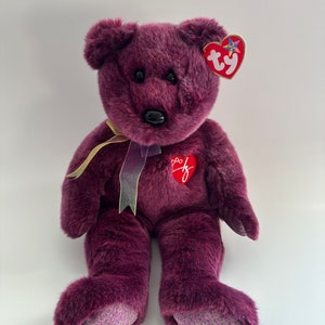 Puede incluir: Oso de peluche morado con una etiqueta de corazón Ty y una cinta morada alrededor del cuello. El oso tiene un corazón rojo en el pecho con un logotipo "Ty".