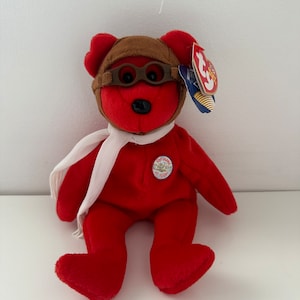 Puede incluir: Oso de peluche rojo de la marca Beanie Baby con un gorro de aviador marrón con gafas y una bufanda blanca. El oso tiene una etiqueta que dice "Ty Beanie Babies" y una etiqueta redonda que dice "Two Worlds of Flight".