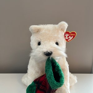 Op de afbeelding: Wit pluche hondenspeelgoed dat een rode en groene sok vasthoudt. De hond heeft een zwarte neus en zwarte ogen. Het speelgoed heeft een Ty Classic-label aan zijn oor.