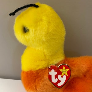 Op de afbeelding: Een gele en oranje gestreepte beanie baby met een zwarte garenantenne. De beanie baby heeft een rood Ty-label met de woorden "Beanie Original Buddy" erop.