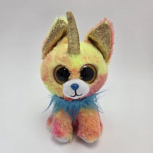 以下が含まれることがあります： 虹色の毛並み、大きな青い目、金色の角を持つ小さなぬいぐるみユニコーン。ユニコーンは青い毛皮の襟をしています。