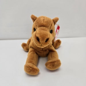 May include: A brown camel plush toy with a red and white tag. The toy is sitting on a white surface.