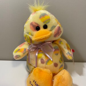 May include: A plush yellow duck toy with a light purple bow and Easter egg patterns. The duck has an orange beak and feet, with the words "Happy Easter" embroidered on one foot. A red tag is attached.