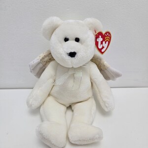 May include: White plush teddy bear with angel wings, wearing a white ribbon. The bear has black eyes and a black nose. The bear has a Ty Beanie Babies tag attached to its right ear.