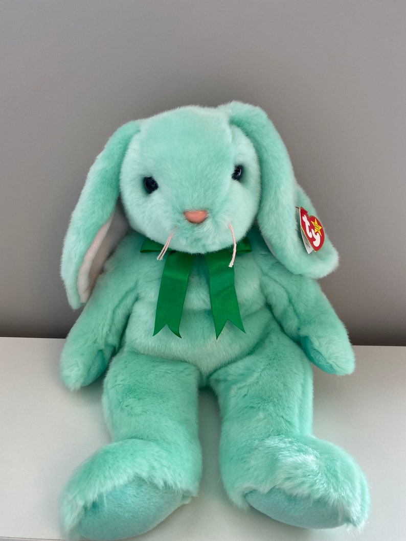 Puede incluir: Un conejo de peluche verde menta con orejas largas y ca&iacute;das y un lazo verde. El conejo tiene una nariz rosa, ojos negros y una etiqueta roja adherida a su oreja. Un peluche suave y tierno.